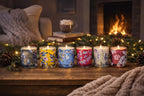 Row of colorful candles on a wooden surface with a fireplace and Christmas decorations in the background.