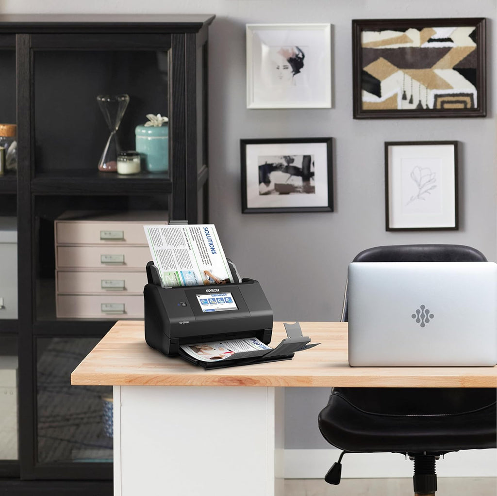 Black document scanner on a desk with a laptop, surrounded by office furniture and decor.