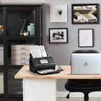 Black document scanner on a desk with a laptop, surrounded by office furniture and decor.