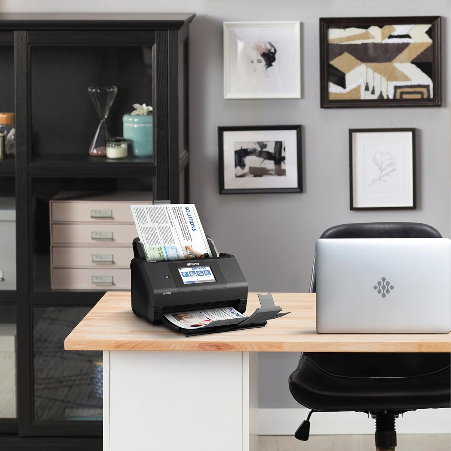 Black document scanner on a desk with a laptop, surrounded by office furniture and decor.