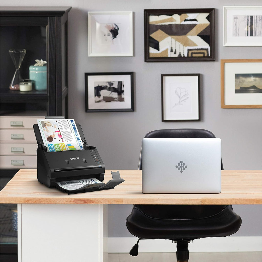 Epson scanner on a desk with a laptop, surrounded by framed pictures on the wall.