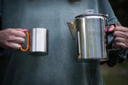 Person holding a metal mug and a metal kettle with a blurred background