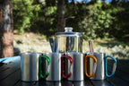 Set of metal camping mugs with colorful carabiner handles on a wooden surface outdoors.