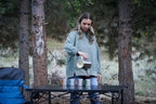 Woman pouring from a metal container into a pot on a camping table in a forest setting