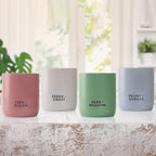 Four scented candles in matte ceramic cups are lined up on a rustic white wooden surface. The candles are in pink, white, sage green, and light blue, with black text labeling their scents: Fern + Bloom, Cedar + Coast, Herb + Meadow, and Velvet + Vanilla.